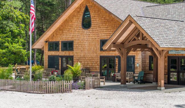 A wooden building with shingle siding, large windows, a covered entrance, and an American flag in front, surrounded by trees, plants, and outdoor seating on a sunny day.