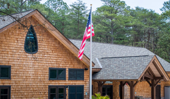 A wooden building with shingle siding and dark blue windows stands in front of a forest of pine trees. An American flag is mounted on a tall flagpole near the entrance.