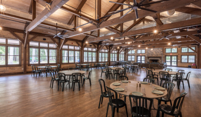 Spacious wooden dining hall with high ceilings, large windows, and exposed beams. Several round tables are set with plates and utensils, and black chairs are arranged around the tables. A stone fireplace is at the far end.