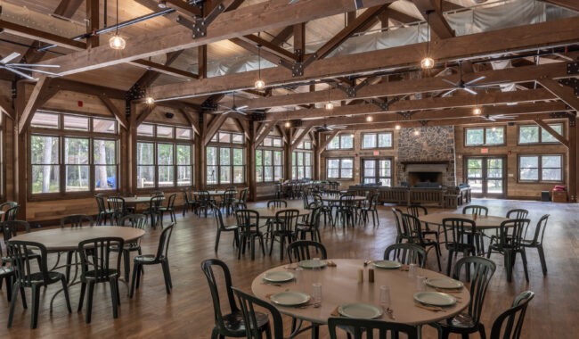 A spacious dining hall with wooden beams, large windows, and a stone fireplace. Several round tables with place settings and black chairs are arranged on a polished wood floor. Natural light fills the room.
