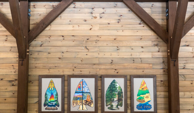 Four framed illustrations of stylized canoe paddles hang on a wooden wall with exposed beams. Each paddle features colorful, nature-inspired designs, and one is labeled Est. 1935.