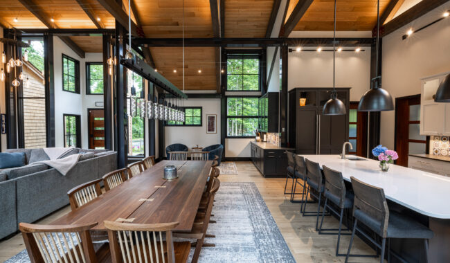 Open-concept kitchen and dining area with high wood ceilings, exposed beams, a long wooden dining table, modern black kitchen cabinets, pendant lighting, and large windows letting in natural light.