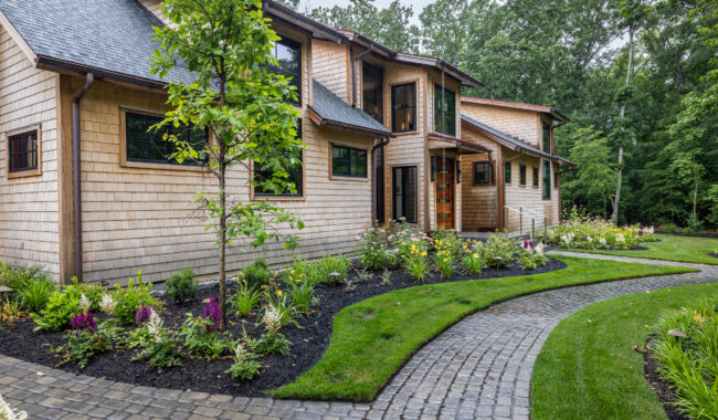 A modern house with tan wood siding, large windows, and a landscaped yard featuring colorful flowers, a curved cobblestone walkway, green grass, and surrounding trees.