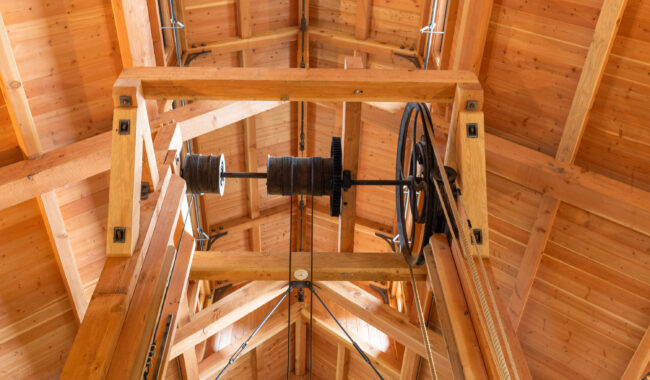 View looking up at a wooden ceiling structure with beams, pulleys, and cables, showing part of a mechanical system suspended from the rafters of a timber-framed building.