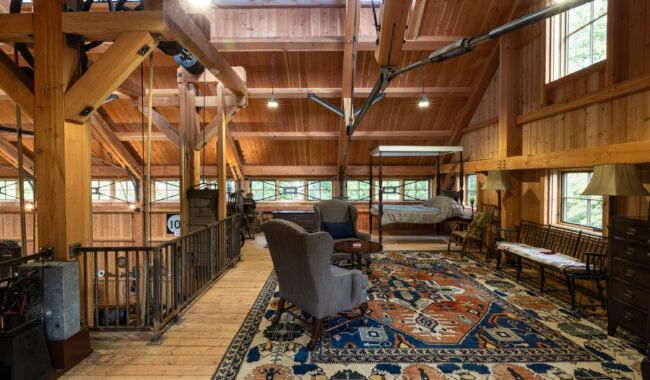 A spacious loft with exposed wooden beams, large windows, a patterned rug, armchairs, a canopy bed, and various lamps and furniture, all bathed in natural light.