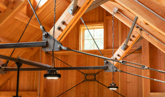 Close-up of exposed wooden beams and metal supports in a vaulted ceiling, featuring industrial hanging lights and a small square window letting in natural light.