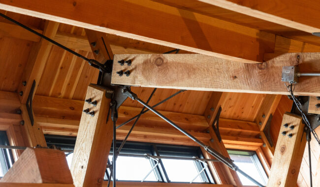 Close-up of wooden beams and metal fasteners in a ceiling structure, with angled support rods and natural light coming through rectangular windows in the background.