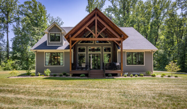 A modern gray house with large windows, a steep gabled roof, and a covered front porch featuring exposed wooden beams, surrounded by grass and tall trees in the background.