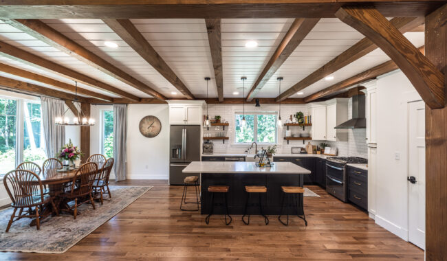 Open-concept kitchen and dining area with exposed wooden beams, white cabinets, stainless steel appliances, a large island with bar stools, and a wooden dining table by large windows overlooking greenery.