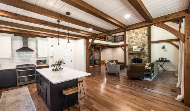 Open-concept kitchen and living area with wooden beams, hardwood floors, white cabinetry, marble island, and a stone fireplace. Cozy seating and natural light enhance the spacious, rustic-modern design.