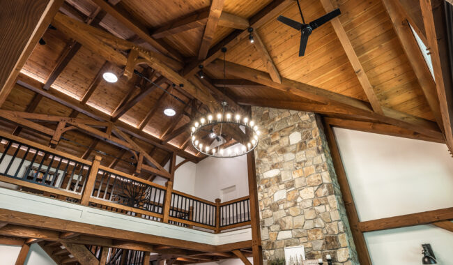 A rustic interior with exposed wooden beams, a high vaulted ceiling, a circular chandelier, a stone fireplace, and a lofted second floor with a wooden railing.
