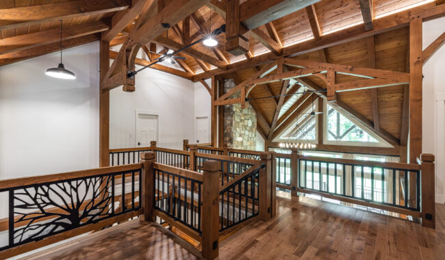 A spacious loft with exposed wooden beams, hardwood floors, and artistic metal railings featuring a tree design. Large windows and a stone wall provide natural light and a rustic, open atmosphere.