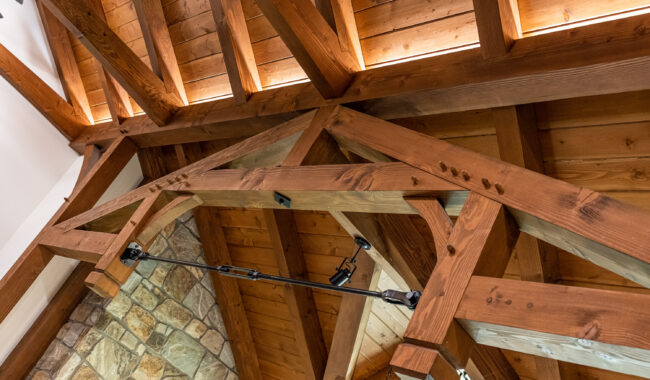 Exposed wooden beams and trusses form the ceiling of a room, with track lighting attached. Part of a stone wall is visible below the warm-toned wood.