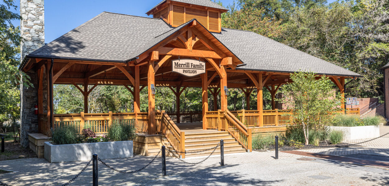 A wooden pavilion labeled “Merrill Family Pavilion” stands surrounded by trees and landscaped bushes, with steps leading up to a covered open-air structure on a sunny day.