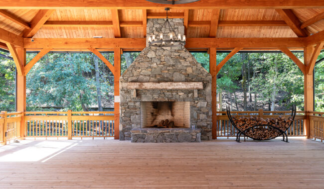 A spacious wooden pavilion with an exposed beam ceiling features a large stone fireplace as the centerpiece, a decorative metal bench to the right, and views of trees through open sides.