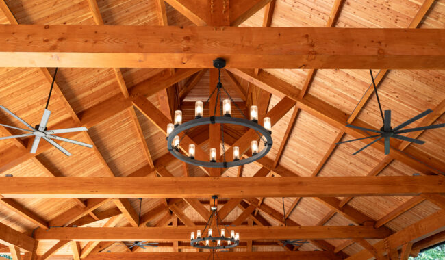 Wooden ceiling with exposed beams, large circular chandeliers with candle-style lights, and two oversized ceiling fans. The structure is spacious and well-lit, with natural wood tones and a geometric pattern.