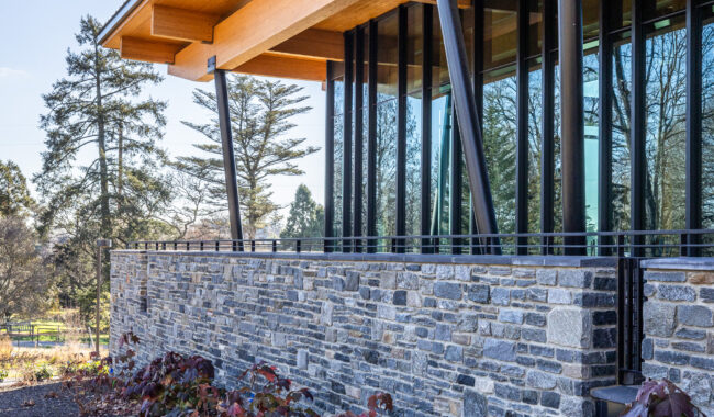 A modern building with a stone facade, large glass windows, and a wooden roof supported by angled black metal columns. Shrubs and bare trees are visible in a landscaped area in the foreground.