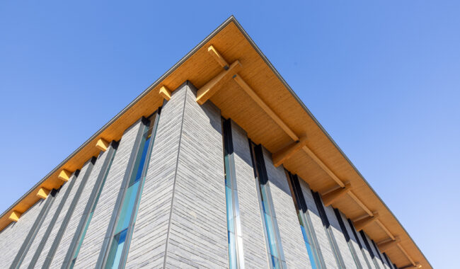 A modern building with tall, narrow windows and a wooden overhanging roof is viewed from below, with a clear blue sky in the background.
