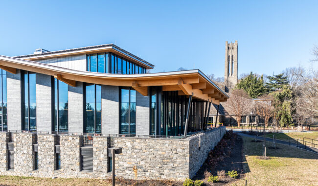Modern building with large glass windows and stone walls, featuring a curved wooden roof. A path leads past the building, with a tall, historic stone tower visible in the background among trees. The sky is clear and blue.