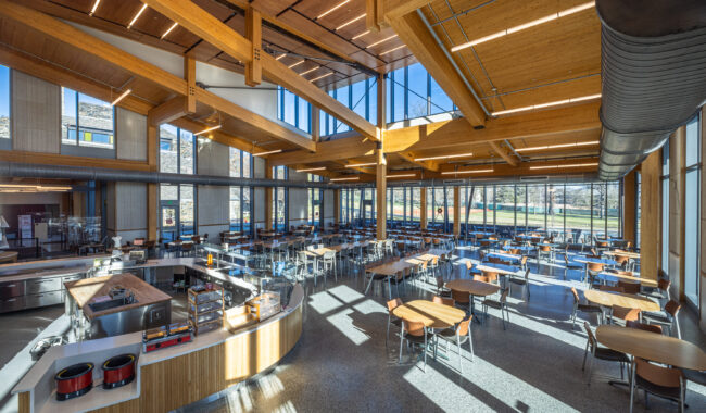 A spacious, modern cafeteria with wooden beams, large windows letting in natural light, rows of tables and chairs, and a central food serving area. Outdoor scenery is visible through the windows.