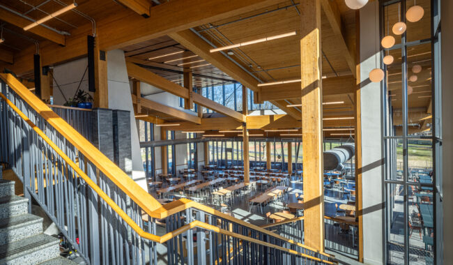 A bright, modern dining hall with wooden beams, large windows, and rows of tables and chairs. A staircase with a wooden handrail is in the foreground, and spherical pendant lights hang from the ceiling.