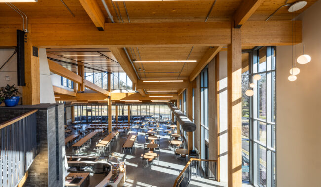 A spacious, modern cafeteria with large windows, wooden beams, and high ceilings. Natural light fills the room, illuminating rows of tables and chairs arranged neatly for dining.