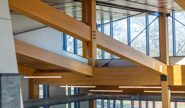 Interior view of a modern building featuring exposed wooden beams, large windows letting in natural light, and visible metal air ducts, with trees visible outside.