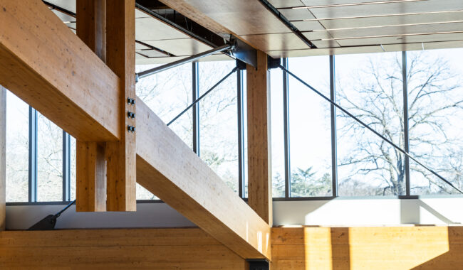 Sunlight fills a modern interior with large wooden beams and posts, metal supports, and tall windows that reveal bare trees and a clear sky outside. The space appears open and architectural.