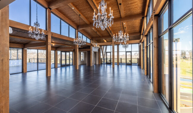 Spacious, modern event hall with large floor-to-ceiling windows, dark tiled flooring, exposed wooden beams, and multiple crystal chandeliers, flooding the room with natural light.