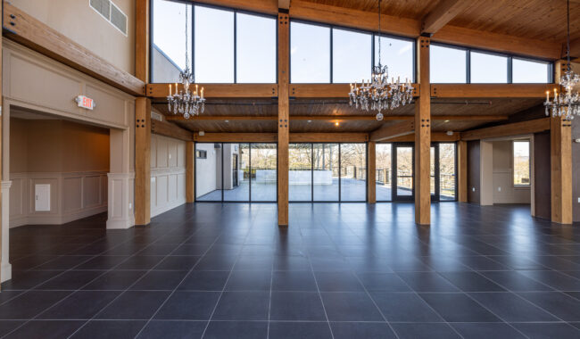 Spacious, empty room with black tile flooring, wooden beams, chandeliers, and large floor-to-ceiling windows providing a view of trees and outdoor area.