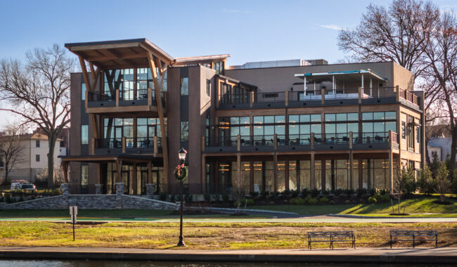 Modern three-story building with large glass windows and balconies, surrounded by trees and greenery, located near a river with benches and a lamppost in the foreground.