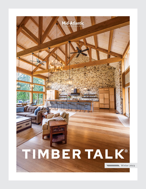 A modern living room with exposed wooden beams, large windows, stone accent wall, a kitchen island, and cozy furniture. The cover text reads Mid-Atlantic Timberframes - Timber Talk, Winter 2023.