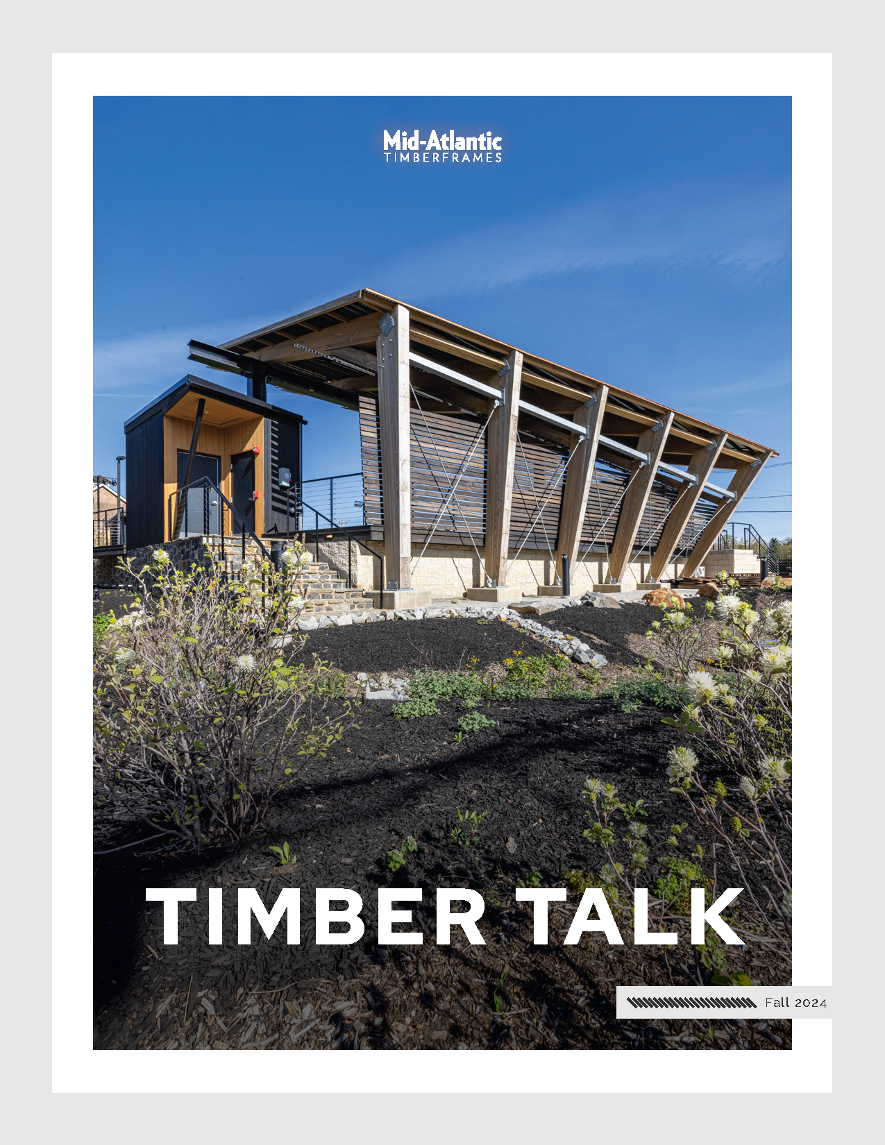 A modern timber-framed building with slanted roof and large windows, surrounded by landscaped plants, under a clear blue sky. Text reads Mid-Atlantic Timberframes, TIMBER TALK, and Fall 2024.