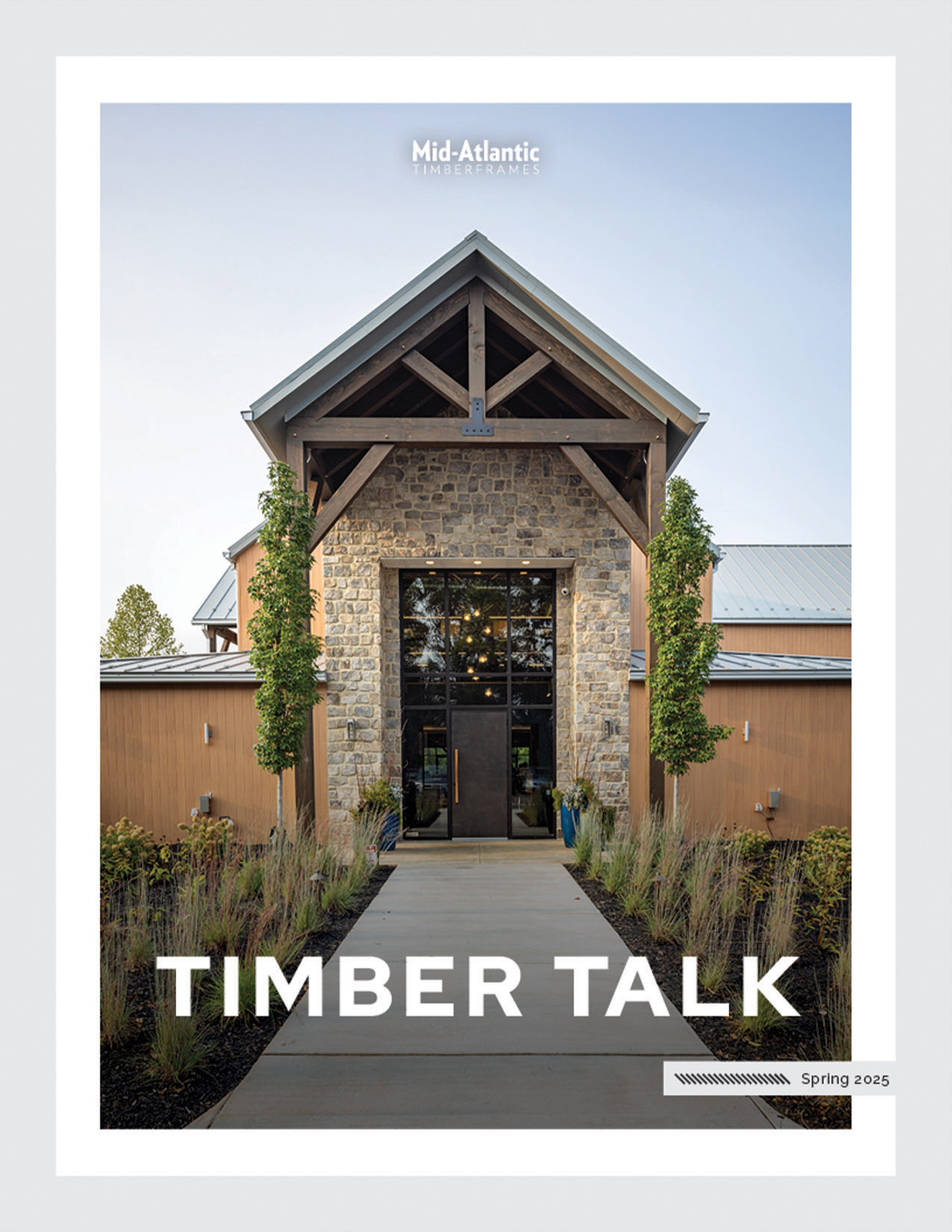 A building with a tall stone entrance and wooden beams is featured on the cover of Timber Talk Spring 2025 by Mid-Atlantic. Lush landscaping lines the walkway leading to the glass front doors.
